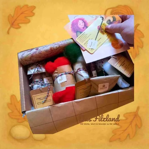 Geöffnete Materialbox, in der eine Hand zwei kleine Filzland-Kärtchen hineinlegt. Sichtbar sind sortierte Wollpäckchen und Zubehör für die Winterprojekte. Persönlicher, handgemachter Eindruck.„Mit Liebe gepackt – von Hand zu Hand.“